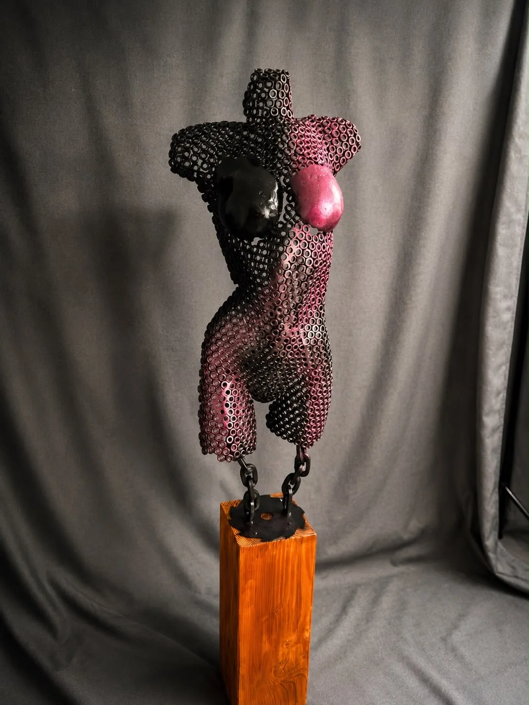Duality — torso van zwarte stalen moeren met roze verloop, aan kettingen boven een houten sokkel